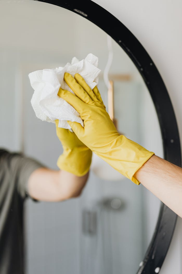 heros-img Close-up of a person cleaning a mirror with yellow gloves, focusing on hygiene and sanitation.